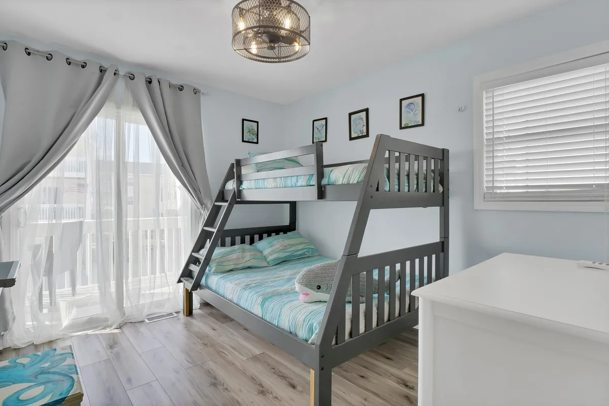 A cheery and inviting bedroom with a twin-over-full bunk bed, white dresser, playful decorative wall art, and a window with sheer curtains letting in abundant daylight. The space is designed for play, relaxation, and family-friendly activities like bedtime stories or games.