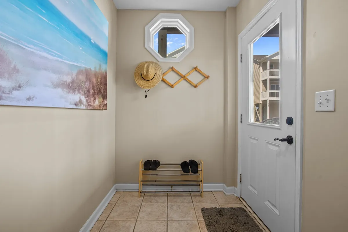 A bright, casual entryway with beach-themed artwork, an octagonal window letting in natural light, and a hat and coat rack with a shoe rack below. The decor sets a beachy, relaxed tone and hints at family-friendly amenities.