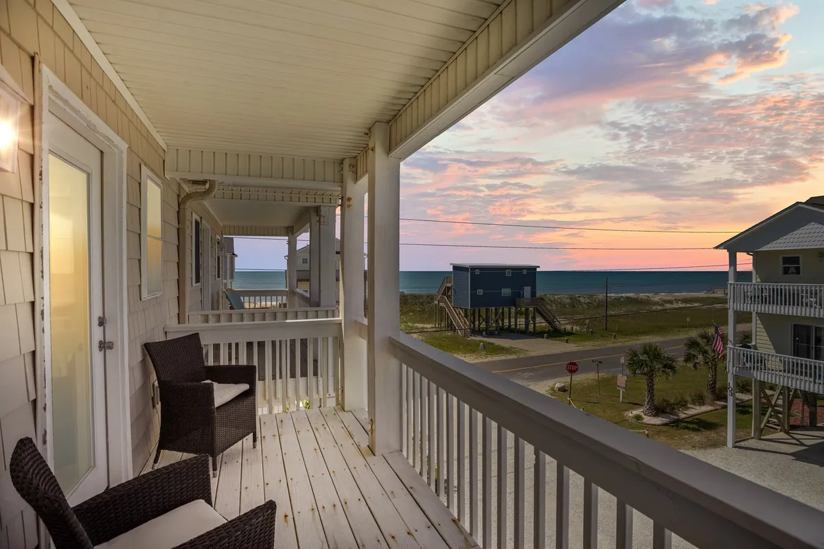 The image showcases the exterior of a charming beach house duplex at dusk, with a spacious balcony overlooking a breathtaking ocean view. The sky is painted with warm sunset hues, and the balcony suggests a perfect spot for relaxing, sipping drinks, and soaking in the tranquil atmosphere while listening to the waves.