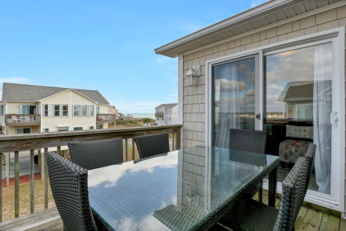 An outdoor glass dining table surrounded by woven chairs sits atop a spacious deck, offering an unobstructed view of the ocean. The setting is both stylish and inviting, suggesting a space ideal for outdoor dining, socializing, or simply soaking in coastal scenery. The comfortable setup reflects the leisurely, communal atmosphere highlighted in the section.