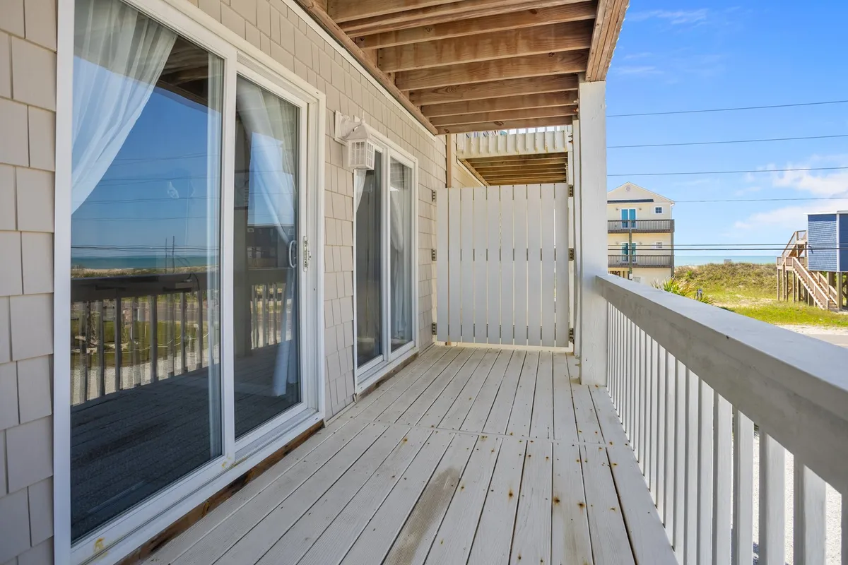 A wooden deck bordered by white railings, furnished with comfortable seating and featuring a clear view of the beach beyond. Sliding glass doors connect the deck directly to the indoor space, blending the inside and outside. The tranquil setting is perfect for enjoying morning coffee, reading a book, or gathering with friends, all with the gentle sound of ocean waves in the background.