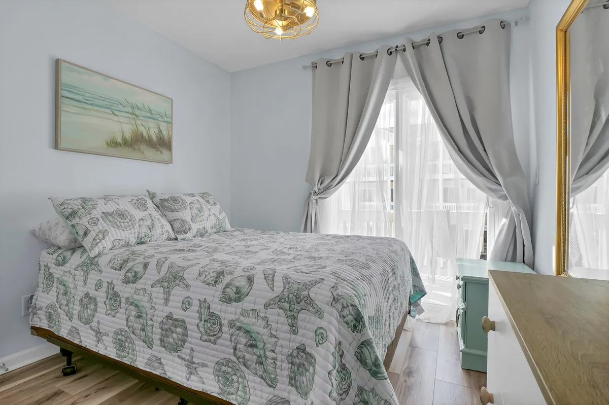 A peaceful bedroom showcasing a bed with a patterned quilt, a large window with curtains that floods the room with natural light, a decorative mirror, and artwork on the wall. The vibe is tranquil and cozy, emphasizing relaxation and comfort.