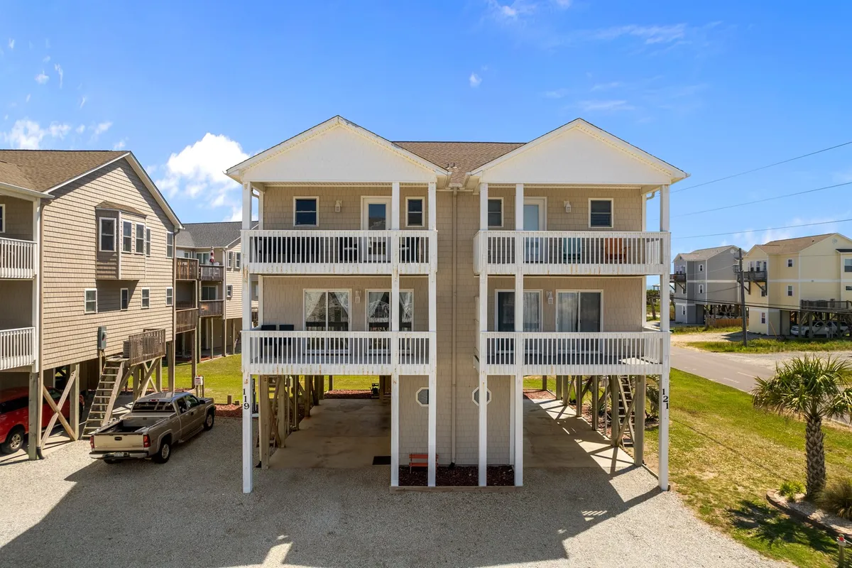 A cheerful, two-story beach house with expansive balconies and surrounding palm trees. The parking area and outdoor spaces highlight the welcoming, family-friendly atmosphere and provide a visual anchor for the duplex mentioned in the text.