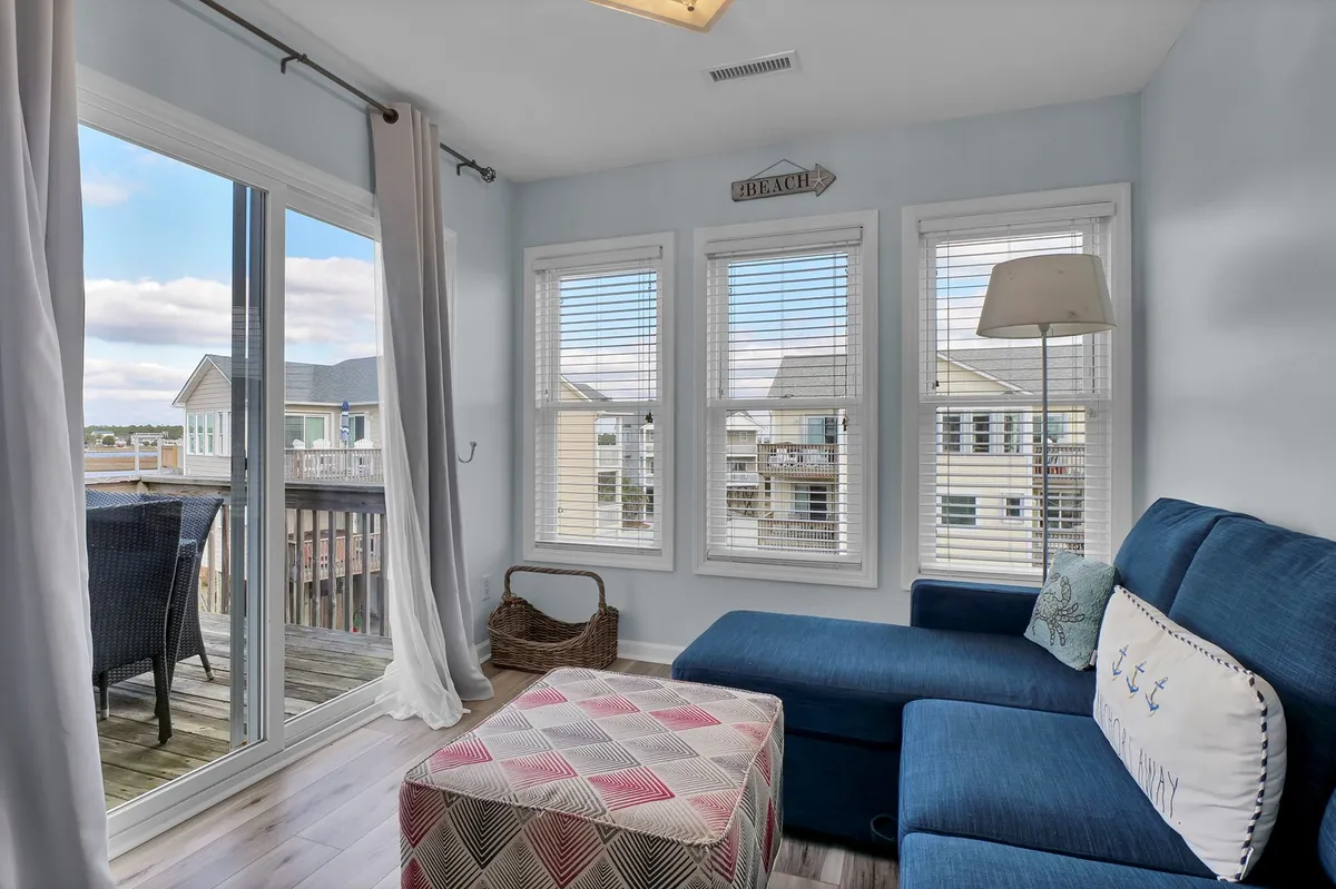 A cozy living space with a blue sectional sofa, colorful ottoman, and large windows that let in ample natural light. The seating is arranged to encourage conversation and relaxation, while the windows offer glimpses of the outdoors and the promise of sunrise views.