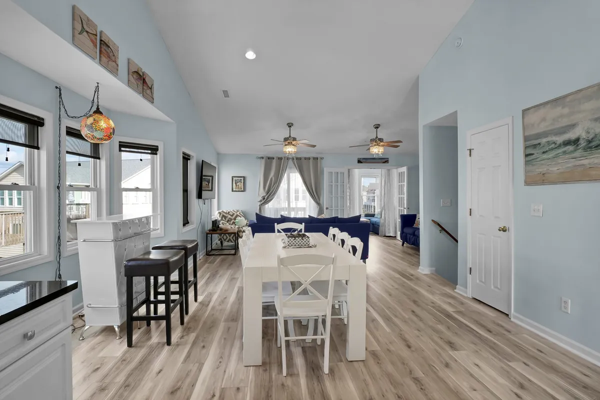 An open, cheerful dining and living area with a dining table, blue sofa, decorative wall art, and abundant natural light. The room feels inviting and suited for family dinners, game nights, and casual group gatherings.