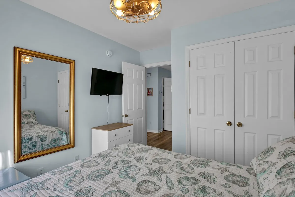 A calming bedroom featuring a bed with intricately patterned bedding, a gold-framed mirror, and a television mounted on the wall. Natural light filters in through a window, creating a soothing atmosphere ideal for relaxing or catching up on favorite shows.