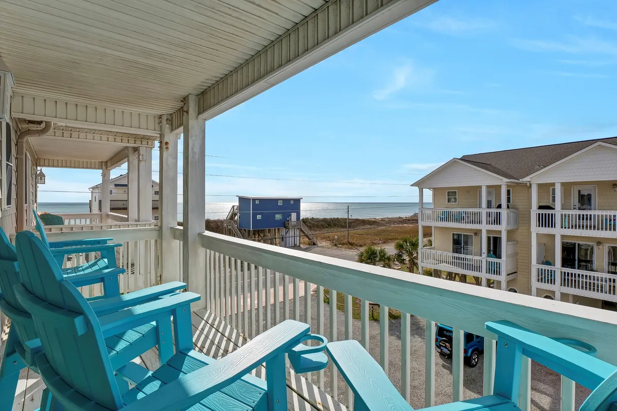 Two turquoise Adirondack chairs are positioned on a deck with a direct ocean view. The setting signals a serene spot for private relaxation, intimate conversations, or simply enjoying the sights and sounds of the sea. The blue house in the distance and fresh sea breeze add to the coastal ambiance.