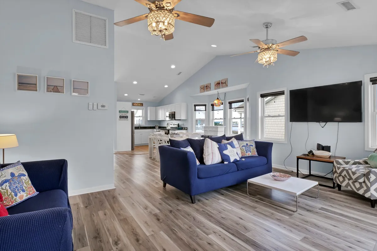 A spacious, open-concept living area featuring a blue sofa, ceiling fans, a smart TV, a dining area, and an adjacent kitchen. Ample windows allow in plenty of natural light, creating a bright and cheerful atmosphere. The space is designed for relaxation, socializing, and effortless flow between living, dining, and kitchen activities.