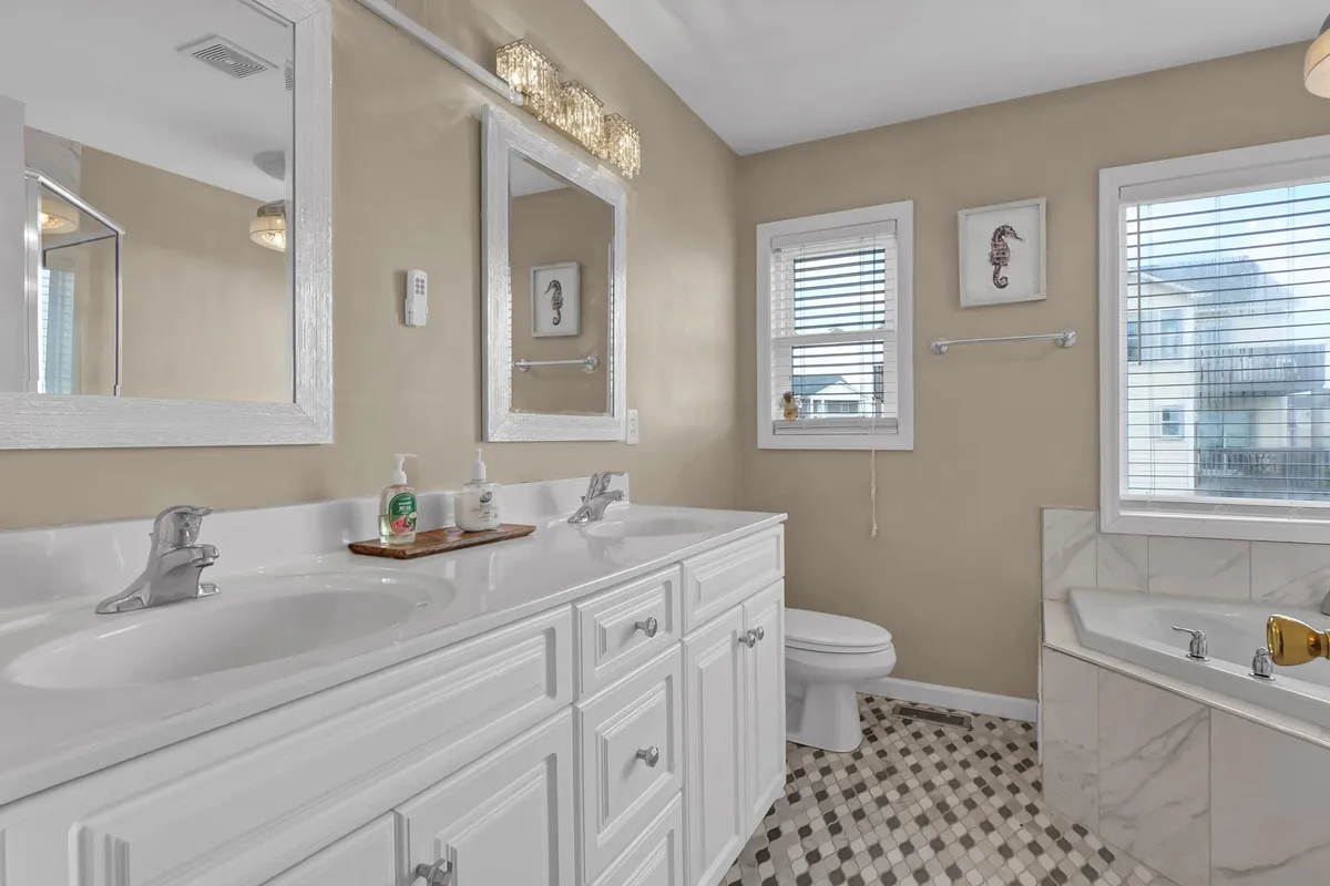 A spa-like bathroom featuring a double sink vanity, large mirrors, a bathtub, and windows that allow for soft natural illumination. The space exudes a refreshing, tranquil mood, ideal for self-care or unwinding after a beach day.