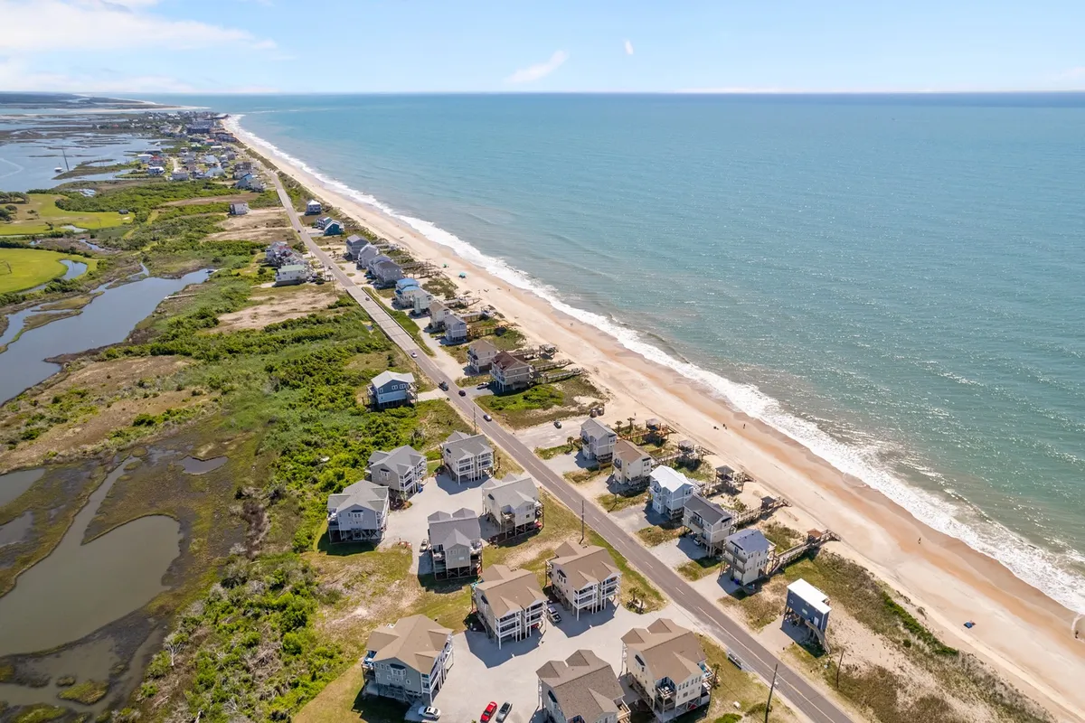 A wide view of beachfront houses, sandy beach, ocean waves, coastal greenery, and a road running alongside the shoreline. The setting is relaxed and serene, with activities like walking on the beach, cycling, birdwatching, and picnicking depicted.