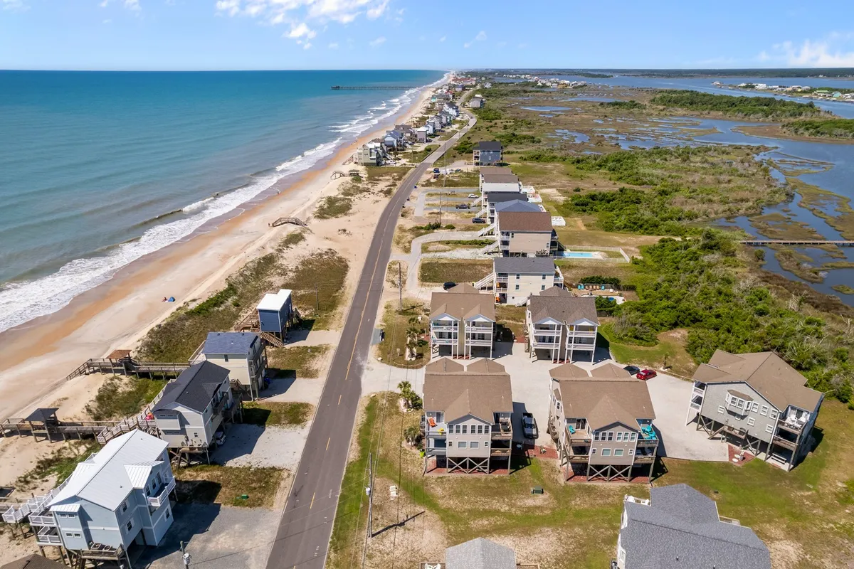 A panoramic viewpoint capturing the ocean, wide sandy beach, and nearby homes, with a road in the foreground. The scene hints at easy access from the property to the shore and offers a sense of the neighborhood vibe and proximity to the water.